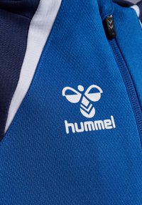 Jacca sportiva blu con tessuto testurizzato, dettagli bianchi e un prominente logo bianco "hummel" con design di ape.