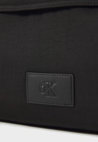 Čierna nylonová taška s textúrovaným povrchom a koženou nášivkou s embosovaným logom "cK", pripevnenou viditeľným prešívaním.