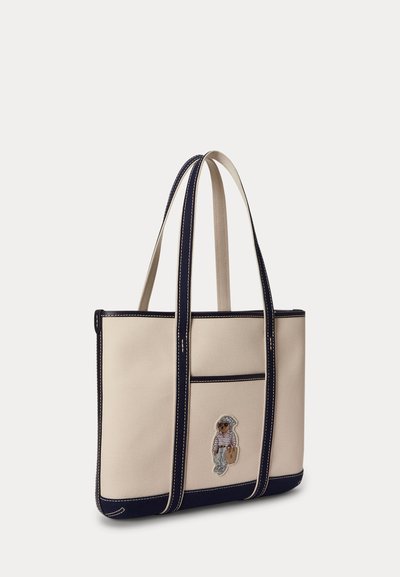 Beige canvas tote bag with navy blue edges and handles, featuring a small embroidered bear wearing casual clothes on the front pocket.