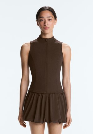 Jeune femme aux cheveux foncés, portant un top marron sans manches ajusté avec une fermeture éclair sur le devant et une mini-jupe évasée, debout devant un fond clair uni.