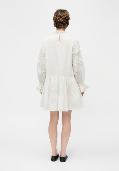 Άτομο με κοντά μαλλιά που φοράει ένα λευκό φόρεμα μακρυμάνικο με λεπτομέρειες δαντέλας και βολάν στο τελείωμα, στέκεται γυρισμένο πίσω, με ορατά μαύρα επίπεδα παπούτσια.