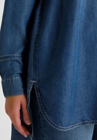 Chemise en jean de couleur bleu foncé, col classique, manches longues et fentes latérales. Présente des surpiqûres visibles et une texture lisse.