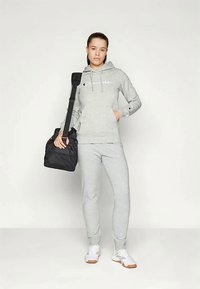 Woman in gray Champion hoodie and sweatpants holding black gym bag over shoulder, standing against plain white background.