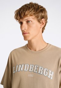 Beige t-paita, jossa on lyhyet hihat ja pyöreä kaula-aukko. Rinnassa on sinisellä ja valkoisella brodeerattu teksti "LINDBERGH 1982".