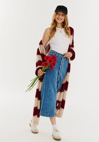 Pruhovaný pletený kardigán v burgundskej a krémovej farbe, biely tielko, džínsová sukňa s vysokým pásom v modrej farbe, drží červené kvety a na nohách má biele tenisky.