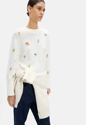 Kobieta w białym dzianinowym swetrze z wielobarwnymi haftowanymi kwiatami, związaną dzianinową odzieżą wokół talii oraz ciemnymi spodniami.