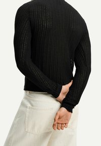 Top de manga larga negro con diseño ajustado, que presenta un patrón de punto texturizado y un cuello redondo, combinado con pantalones de color claro.