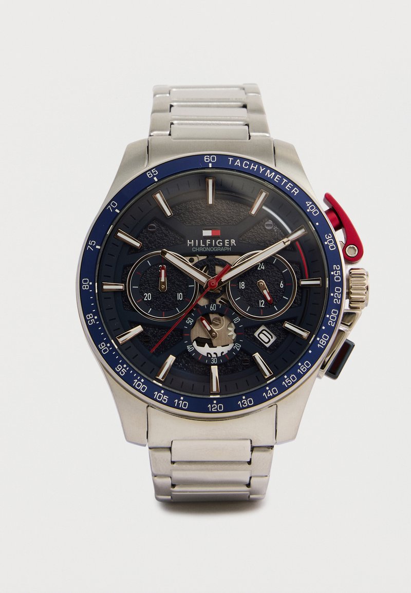 Ασημένιο μεταλλικό ρολόι χρονογράφου με μπλε στεφάνη ταχυμέτρου, μαύρο καντράν, τρία υποκαντράν, κόκκινους και χρυσούς δείκτες και μεταλλικό λουράκι με συνδέσμους.
