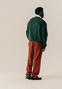 Mann mit kurzen lockigen Haaren, der einen grünen Pullover, rote Hosen, schwarze Schuhe und eine goldene Brille trägt, steht seitlich vor einem beigen Hintergrund.