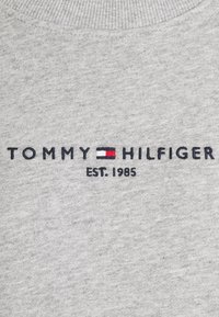 Grå sweatshirt med ribbad halsringning. Har broderad "TOMMY HILFIGER"-logotyp i marinblått med röda och vita detaljer. Grundades 1985.