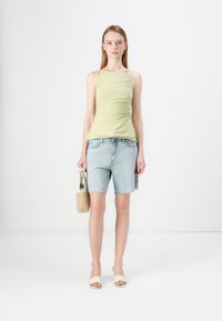 Lichtgroene mouwloze top met ruches, gecombineerd met denimshorts. Model houdt een geweven tan tas vast en draagt lichte sandalen. Eenvoudig, minimalistisch ontwerp.