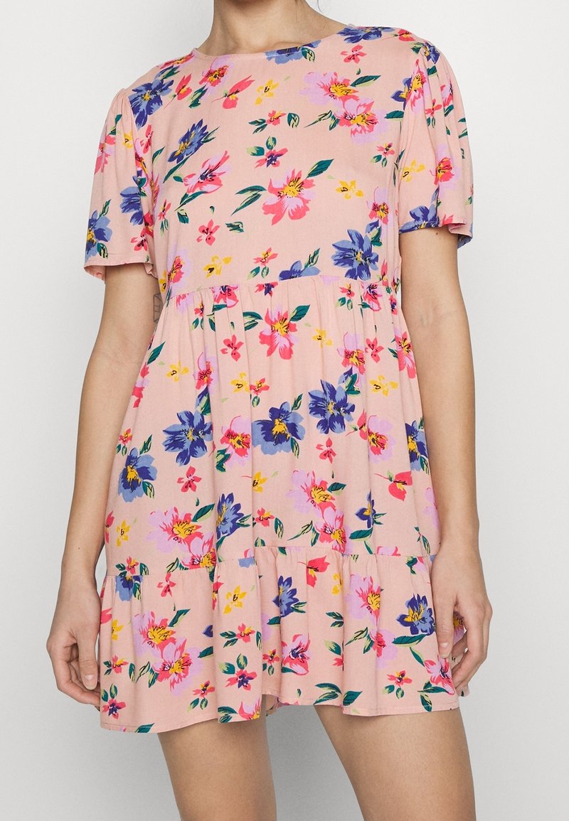 Femme portant une courte robe rose à imprimé floral multicolore et manches courtes, debout les bras détendus le long du corps.