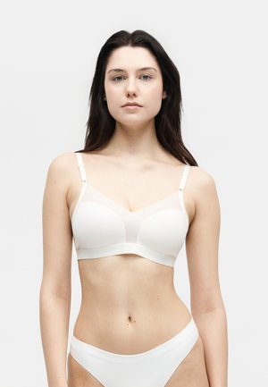 Kobieta o długich ciemnych włosach, ubrana w biały bralette i dopasowaną białą bieliznę, stojąca na jasnym, jednolitym tle.