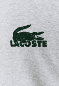Harmaa puuvillakangas, jossa on vihreä krokotiililogo ja rohkealla fontilla teksti "LACOSTE", näyttäen sileän rakenteen.