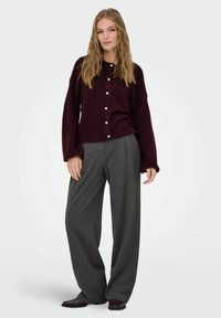 Burgunderfarbener Strick-Cardigan mit rundem Ausschnitt und weißen Knöpfen, kombiniert mit grauen, weiten Hosen und dunklen Schuhen, getragen von einem stehenden Model.