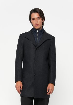 Homme portant un manteau en laine gris foncé à boutons sur une veste zippée bleu marine, se tenant devant un fond blanc uni.