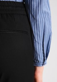 Pantaloni neri con una texture liscia, dotati di tasca laterale, abbinati a una camicia a righe blu e bianche con polsini abbottonati e maniche arrotolate.