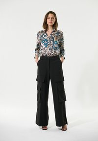 Geprinte blouse met blauw en beige patroon, gecombineerd met zwarte cargo-broek met meerdere zakken en zijslitsen. Schoenen zijn zwarte hoge hakken.
