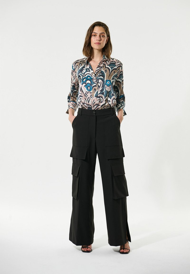Geprinte blouse met blauw en beige patroon, gecombineerd met zwarte cargo-broek met meerdere zakken en zijslitsen. Schoenen zijn zwarte hoge hakken.