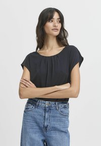 Schwarze kurzärmelige Bluse mit rundem Halsausschnitt und gefältetem Detail, kombiniert mit hochtaillierten blauen Jeans.