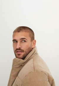 Giacca in corduroy beige con colletto alto, caratterizzata da un motivo a coste testurizzate e chiusura con zip, indossata da un uomo con capelli corti.
