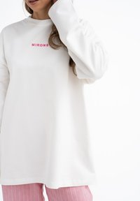 Wit T-shirt met lange mouwen en roze opdruk "MIRONS". Zacht materiaal, relaxte pasvorm en ronde hals. Gecombineerd met roze gestreepte broek.
