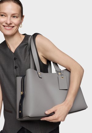 Mujer sonriente con chaleco oscuro sin mangas sostiene un gran bolso tote de cuero gris con herrajes dorados y detalle de cremallera negra sobre un fondo liso.