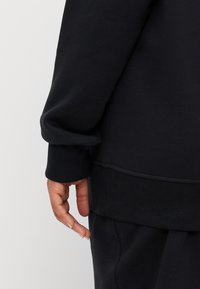 Svart hettegenser med myk tekstur, med en avslappet passform. Mansjetter og hem er ribbet. Hånd vist hvilende nær plaggets side.