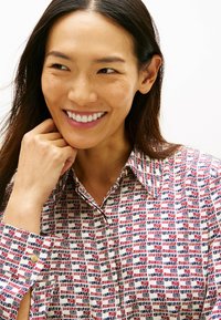 Camisa con cuello que presenta un patrón multicolor en rojo, azul y blanco. La tela parece ligera con una textura suave. Detalle de botón dorado en el puño.