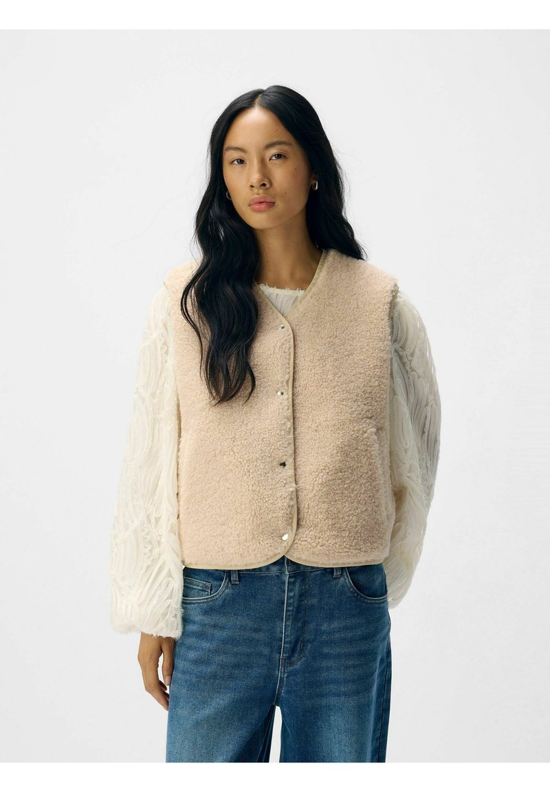 Lys beige fleece vest med rund nederste kant og trykknapper, båret over en struktureret hvid langærmet top, parret med blå jeans.