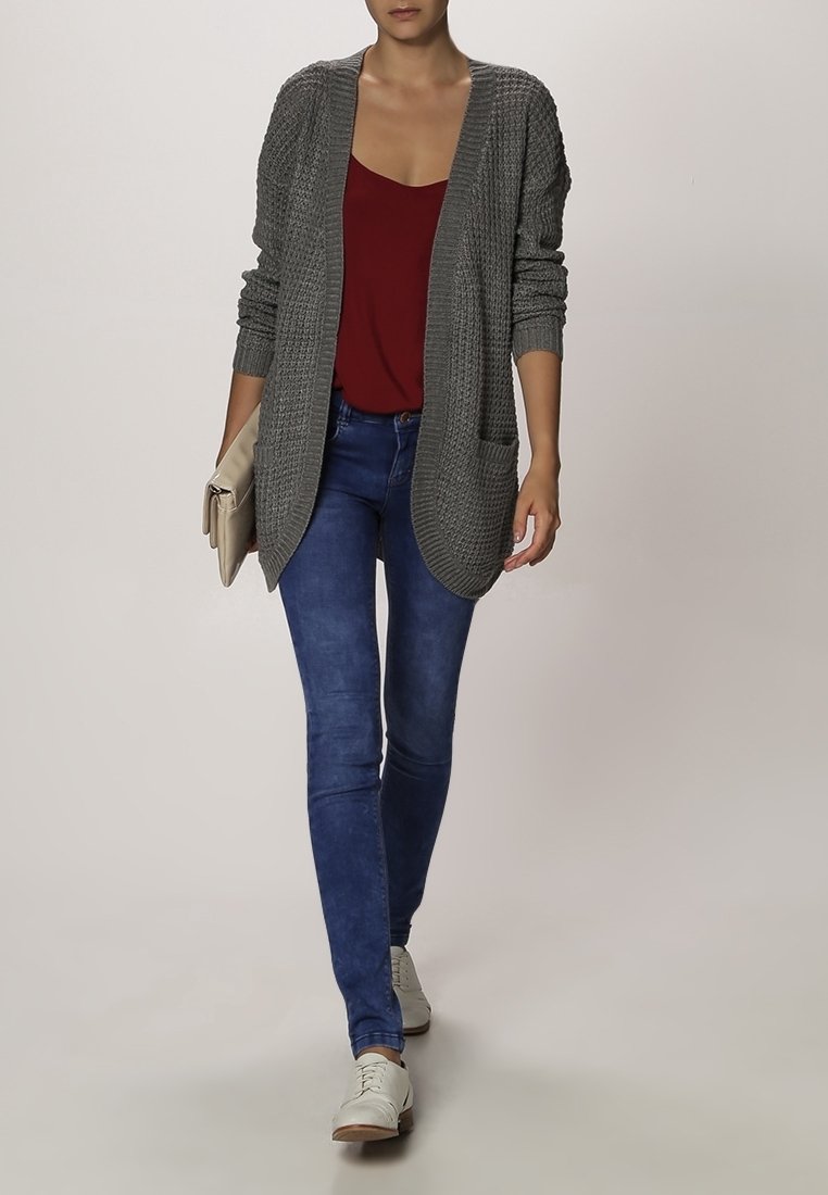 Femme marchant portant un cardigan gris tricoté, un haut bordeaux, un jean bleu, des chaussures blanches et tenant une pochette beige.