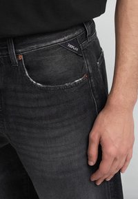 Jeans in denim nero con una texture sbiadita, caratterizzati da un distintivo dettaglio della tasca sfrangiato e un'etichetta "REPLAY" sulla vita.