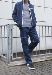 Modrá fleecová bunda s bílým zipem, šedý pletený svetr, tmavé denimové kalhoty s panelovým prošíváním a černé tenisky s bílou podrážkou.