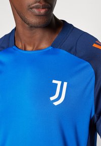 Camisa atlética azul com detalhes em azul escuro, apresentando um logótipo branco. Textura suave e detalhes de decote redondo visíveis, realçando o contraste de cores.