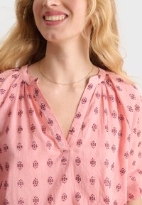 Blusa di cotone rosa con scollo a V, caratterizzata da un motivo floreale in rosa scuro. Design plissettato attorno allo scollo e maniche corte.