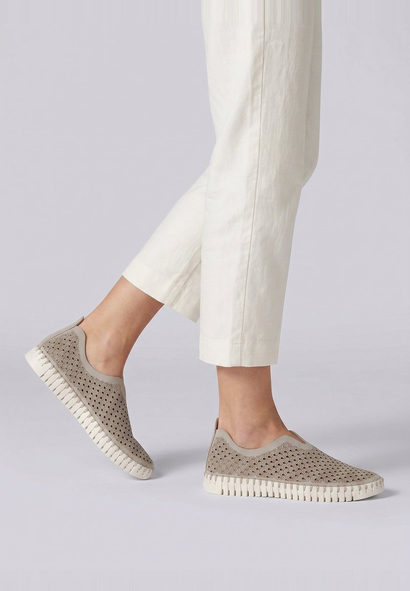 Persona que lleva zapatos beige tipo slip-on con patrón de ondas y suelas blancas, combinados con pantalones blancos recortados contra un fondo liso.