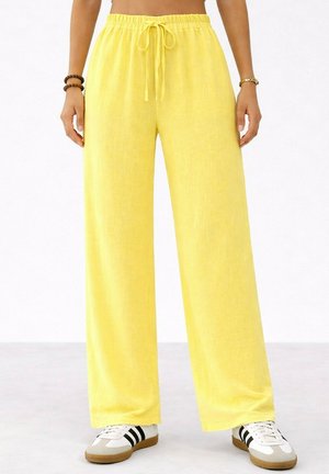 Pantalon jaune à jambes larges avec taille à cordon, porté avec des baskets blanches à rayures noires sur un sol gris clair.