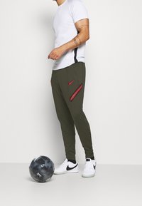 Gröna sportbyxor har en röd Nike-logotyp och sidofickor med röda och teal-randiga detaljer. Den vita t-shirten är ihopparad med svartvita sneakers.