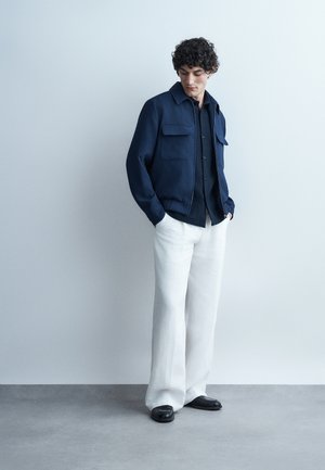 Άνδρας με σγουρά μαλλιά, φοράει σκούρο μπλε σακάκι και πουκάμισο, άσπρο παντελόνι με φαρδιά πόδια και μαύρα παπούτσια, στέκεται μπροστά σε γκρι ανοιχτό τοίχο.