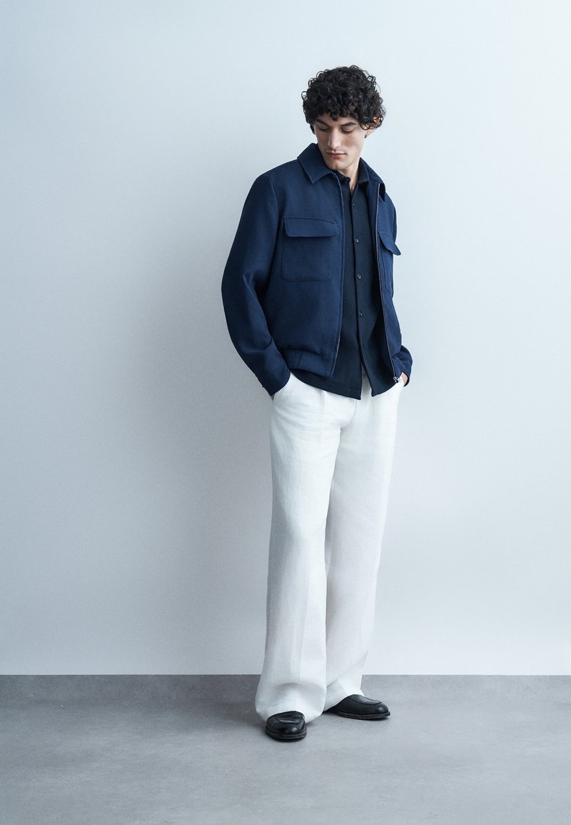 Άνδρας με σγουρά μαλλιά, φοράει σκούρο μπλε σακάκι και πουκάμισο, άσπρο παντελόνι με φαρδιά πόδια και μαύρα παπούτσια, στέκεται μπροστά σε γκρι ανοιχτό τοίχο.