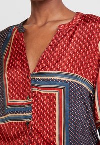 Chemise rouge à motifs avec un col en V, présentant des dessins géométriques en bleu et crème, tissu texturé, et boutons décoratifs près de l'encolure.