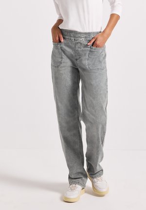 Osoba nosící šedé kalhoty s elastickým pasem a rovnými nohavicemi, s předními našitými kapsami a bílými teniskami, stojící na jednobarevném bílém pozadí.