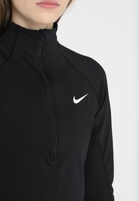 Musta vetoketjullinen paita, joka on valmistettu joustavasta kankaasta. Tässä paidassa on korkea kaulus, ribbireunat ja pieni valkoinen Nike-logo vasemmassa rintapielessä.