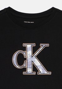 Camiseta de algodón negra con un gran logo "CK" en un patrón de nubes azules y blancas, contorneado en beige delgado, con un diseño clásico de cuello redondo.