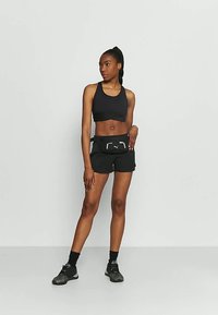 Black sports bra and black shorts, accompanied by a fanny pack with a logo. Model wears black socks and athletic shoes, standing against a neutral backdrop.