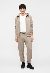 Beige hoodie en bijpassende joggingbroek van zachte stof, met een trekkoord in de taille en zijzakken, gecombineerd met zwarte sneakers.
