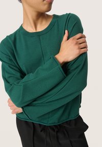Groene pullovertrui van zachte stof met een ronde halslijn, verlaagde schouders en lange mouwen; gecombineerd met zwarte broek met koord in de taille.