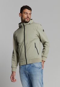 Licht olijfgroene ritsjack met capuchon, voorzien van een zwarte rits en zakken. Glad stof en ribgebreide manchetten, gedragen over blauwe jeans.