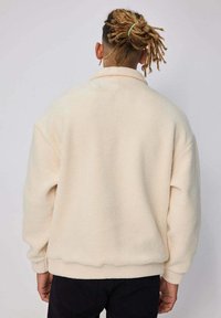 Pull à col montant en polaire de couleur crème, avec une coupe décontractée et des poignets côtelés. Texture douce, design minimal et sans marquage visible.