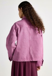 Růžová džínová bunda s volnějším střihem, viditelným prošitím a knoflíkovými manžetami. Hladká textura s mírně vybledlým finišem na látce.